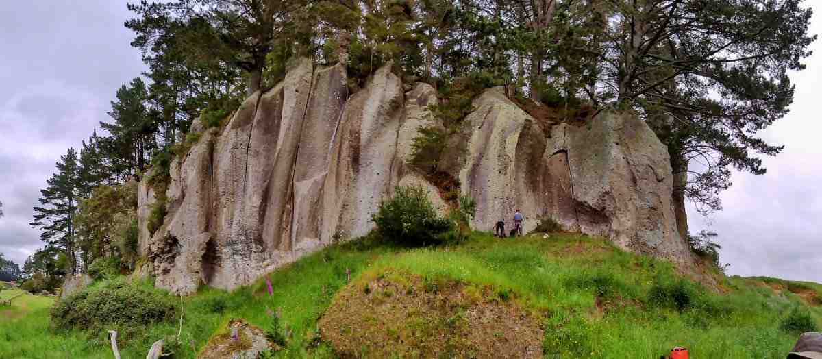 Wide angle view of Bayley Road ignimbrite rock crag topped by pine trees with climbers at base