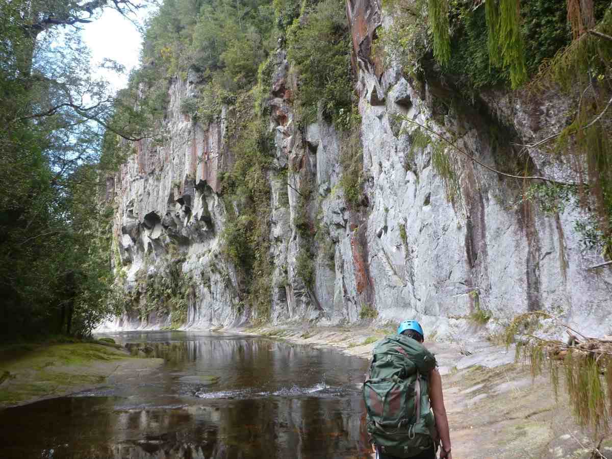 Hiker with pack approaches a cliff reflected in Mangorewa river
