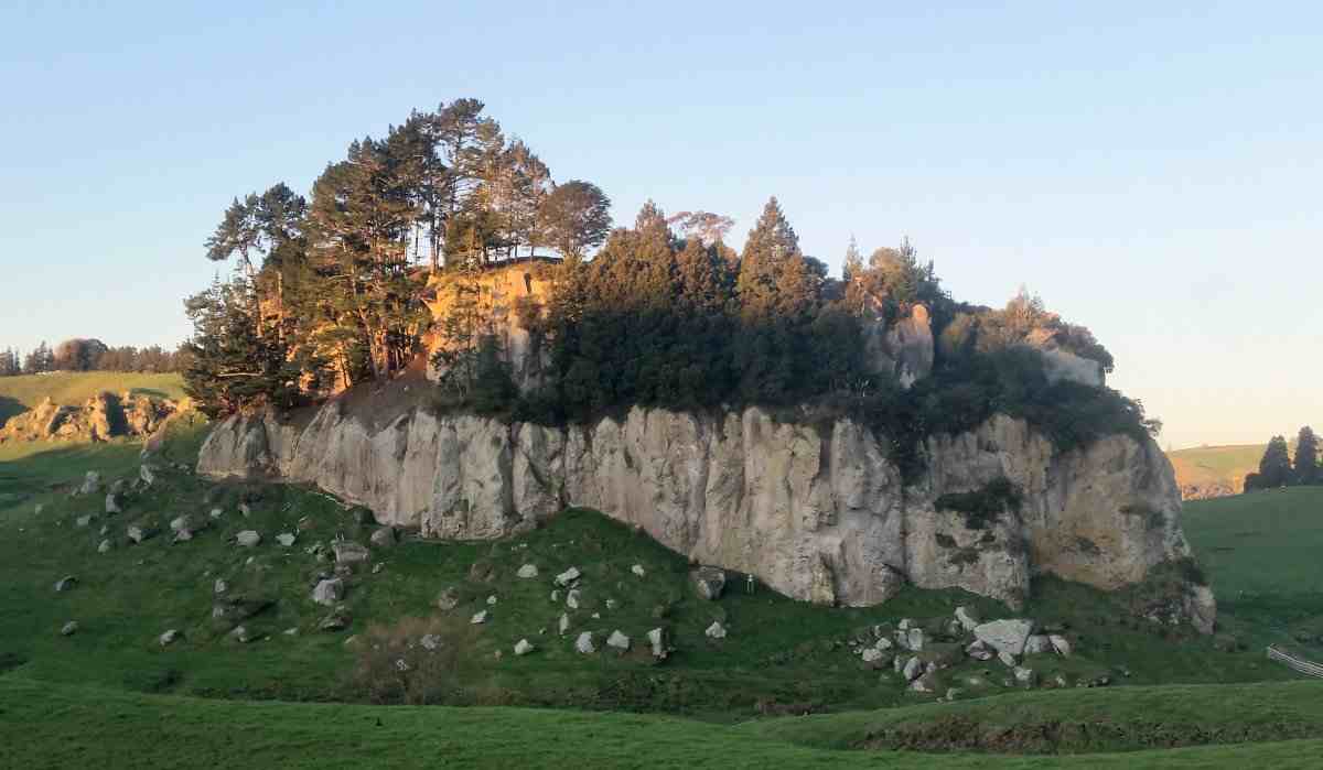 Sun shining on the top of a rock plug rising from green farmland topped by pine trees.