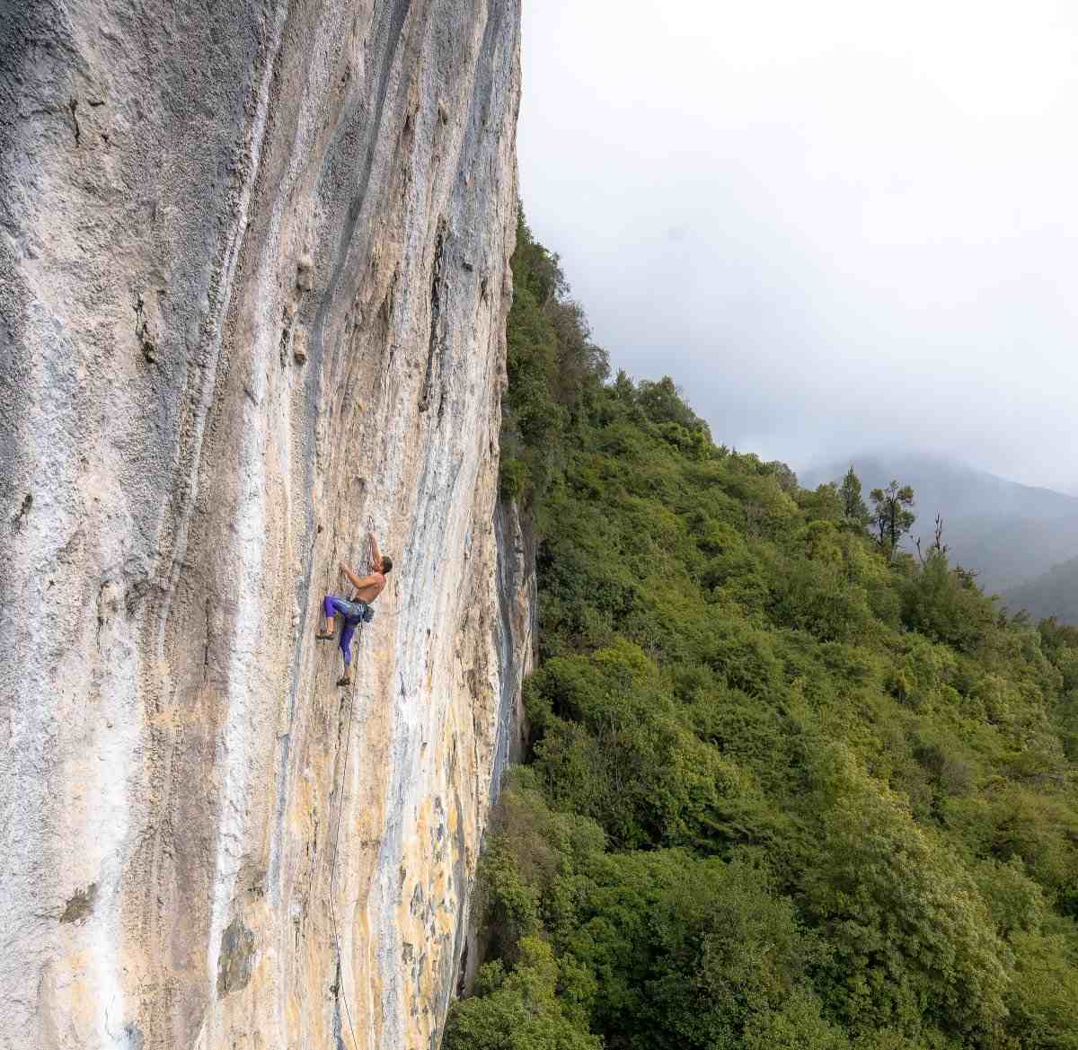Climber on overhanging orange white rock above bush