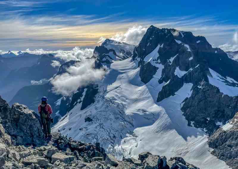 Person stands on a rocky ridge looking away from the camera but towards a glacier and large snowy mountains in front of them