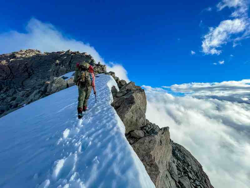 Person walking uphill on snowy ridge, blue sky and clouds in background