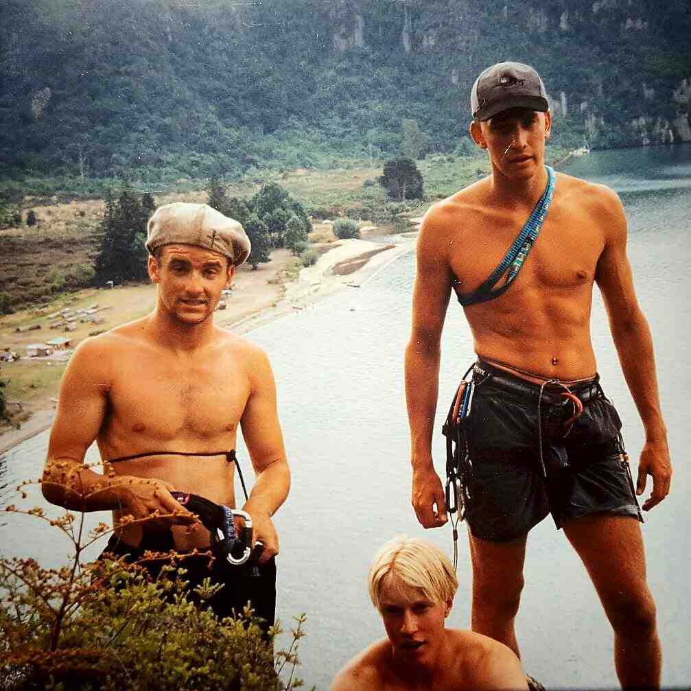 Three climbers with shirts off, 2 wearing hats, high above a lake and beach in background.