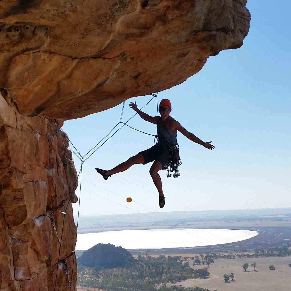 Smiling climber hangs under an orange rock roof with a lake in the background.