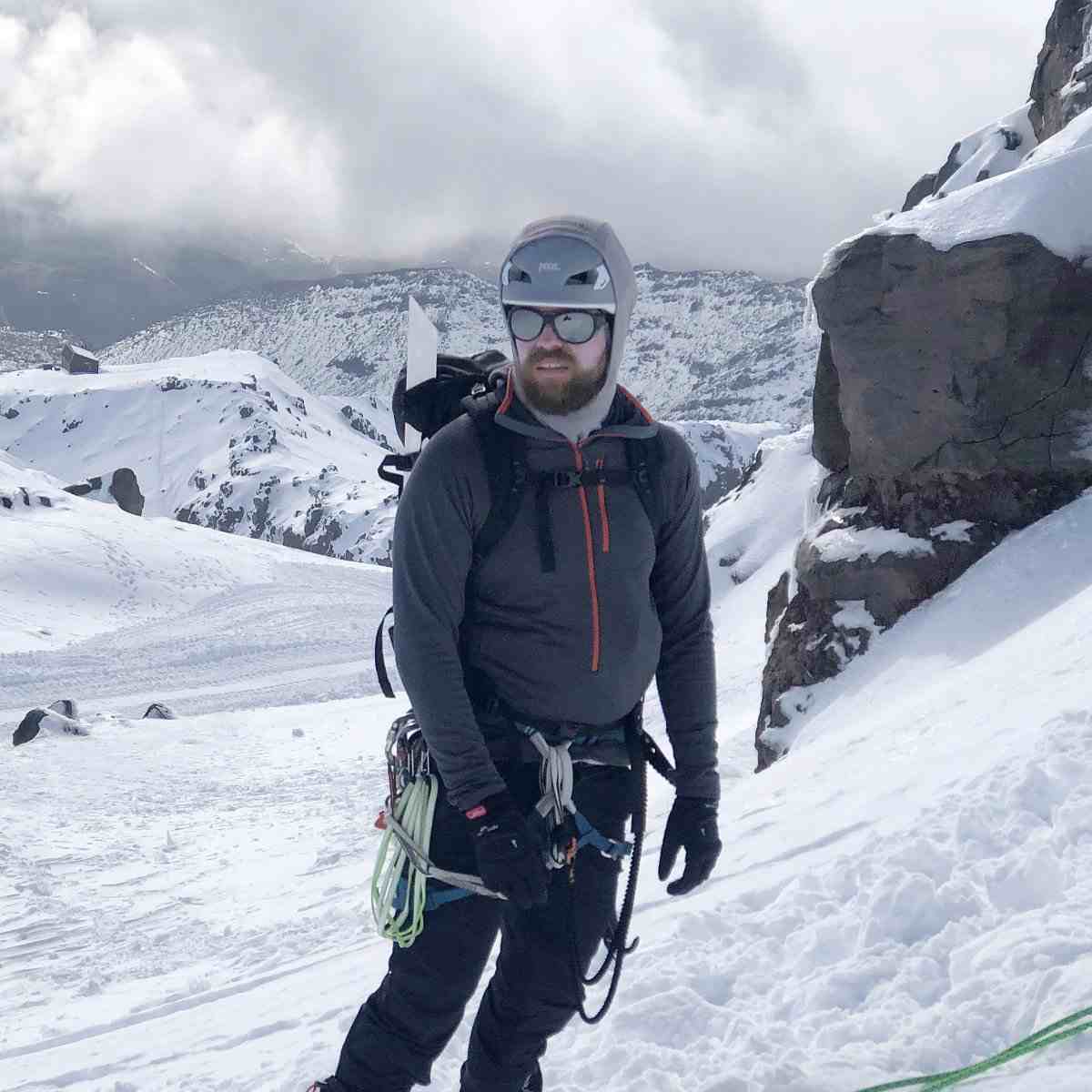 Bearded and helmeted climber in sunglasses with alpine equipment and snowy background