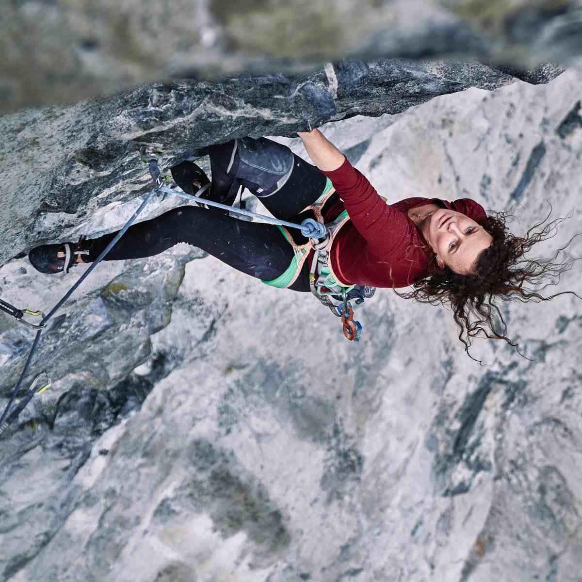 Female climber in red shirt with wild hair looking up on steeply overhanging rock.
