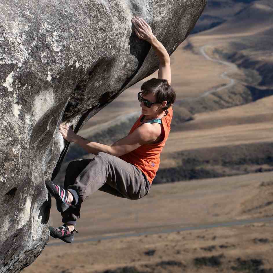 Female climber in sunglasses and orange top on sloping overhanging boulder.