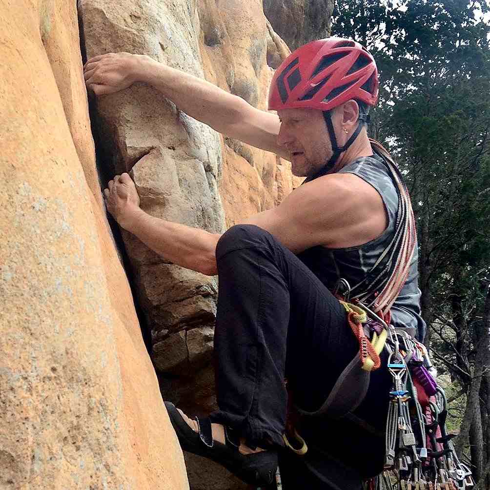 Climber in red helmet with bare arms and a lot of gear on harness high-steps up a crack on orange rock.