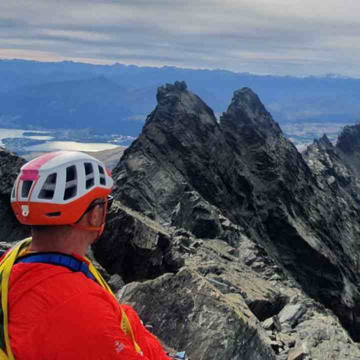 Climber in red top and red/white helmet looks at twin sharp peaks high above a lake.