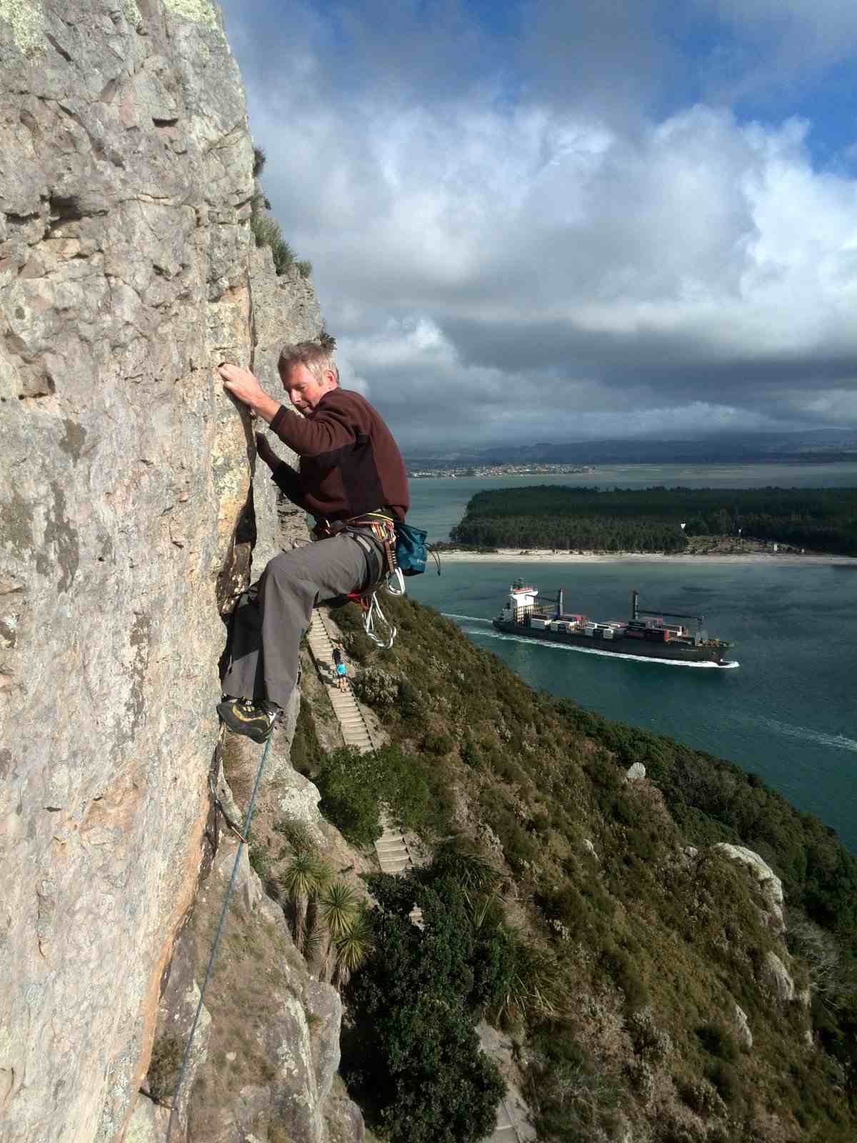 Climber without helmet on slightly overhanging rock face above a harbour with container ship.