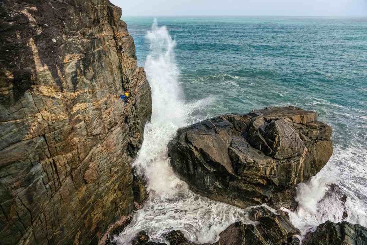 Tiny climbers on a brown rock sea cliff with pounding waves and towering spray