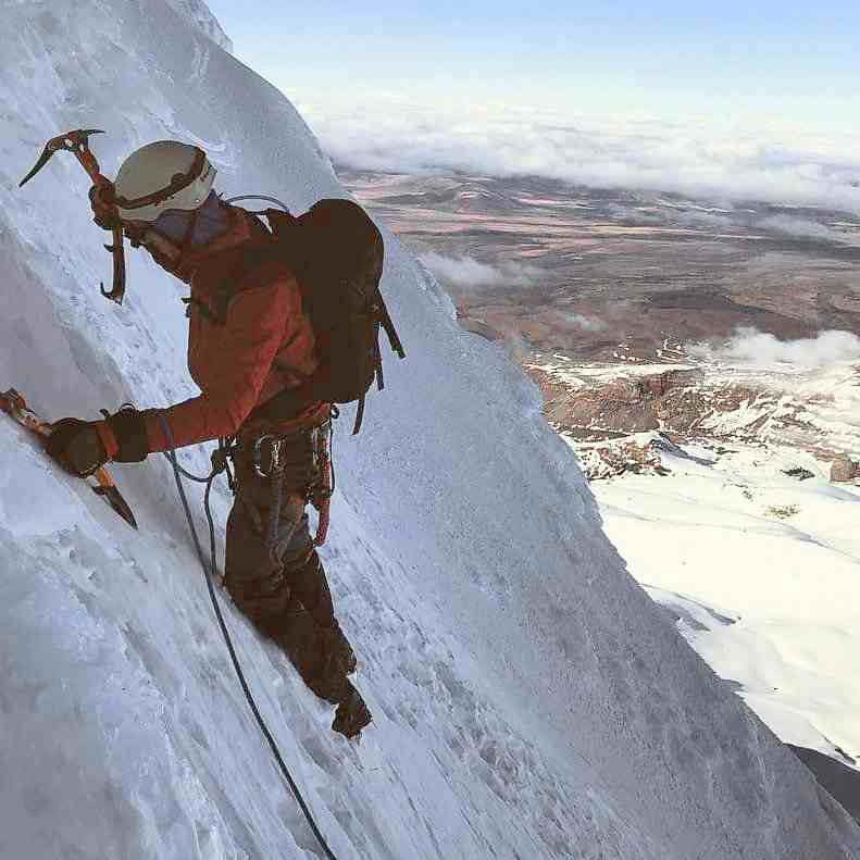 Climber with ice axes, red jacket and white helmet on a steep snow slope in shadow