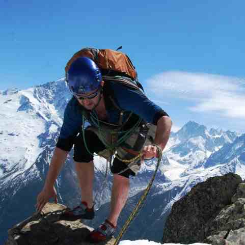 Climber wearing shorts and a red pack on a rocky peak with snow covered mountains behind under a blue sky.