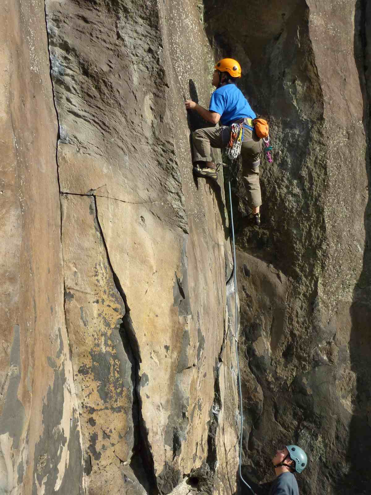 Climber in blue shirt and orange helmet on warm-colored rock corner with belayer looking up.