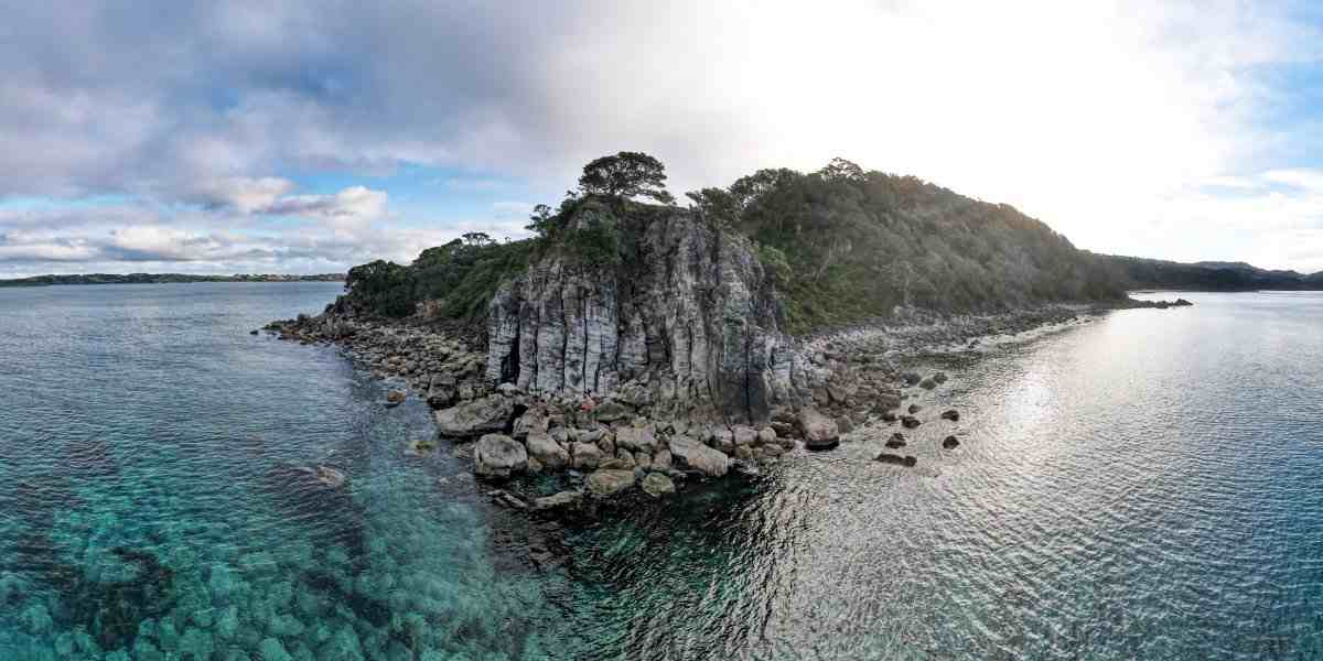 Wide angle aerial image of Ti Point, basalt headland with pohutakawa, calm turquoise sea, underwater reef, cloudy sky.