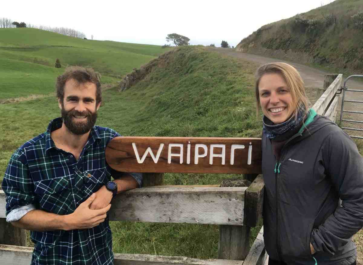 Bearded man and blonde woman either side of a Waipari sign in front of farmland