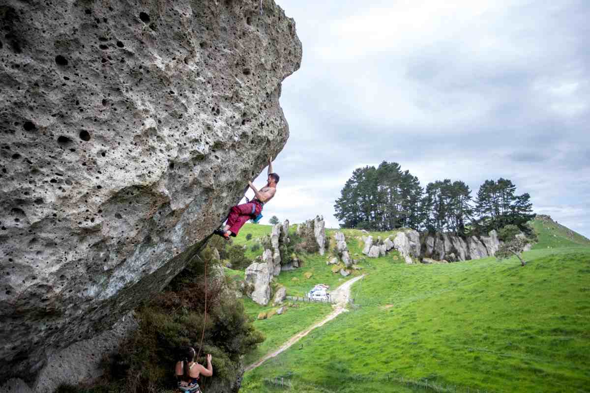 Shirtless climber in red trousers on steeply overhanging pocketed rock with farmland background