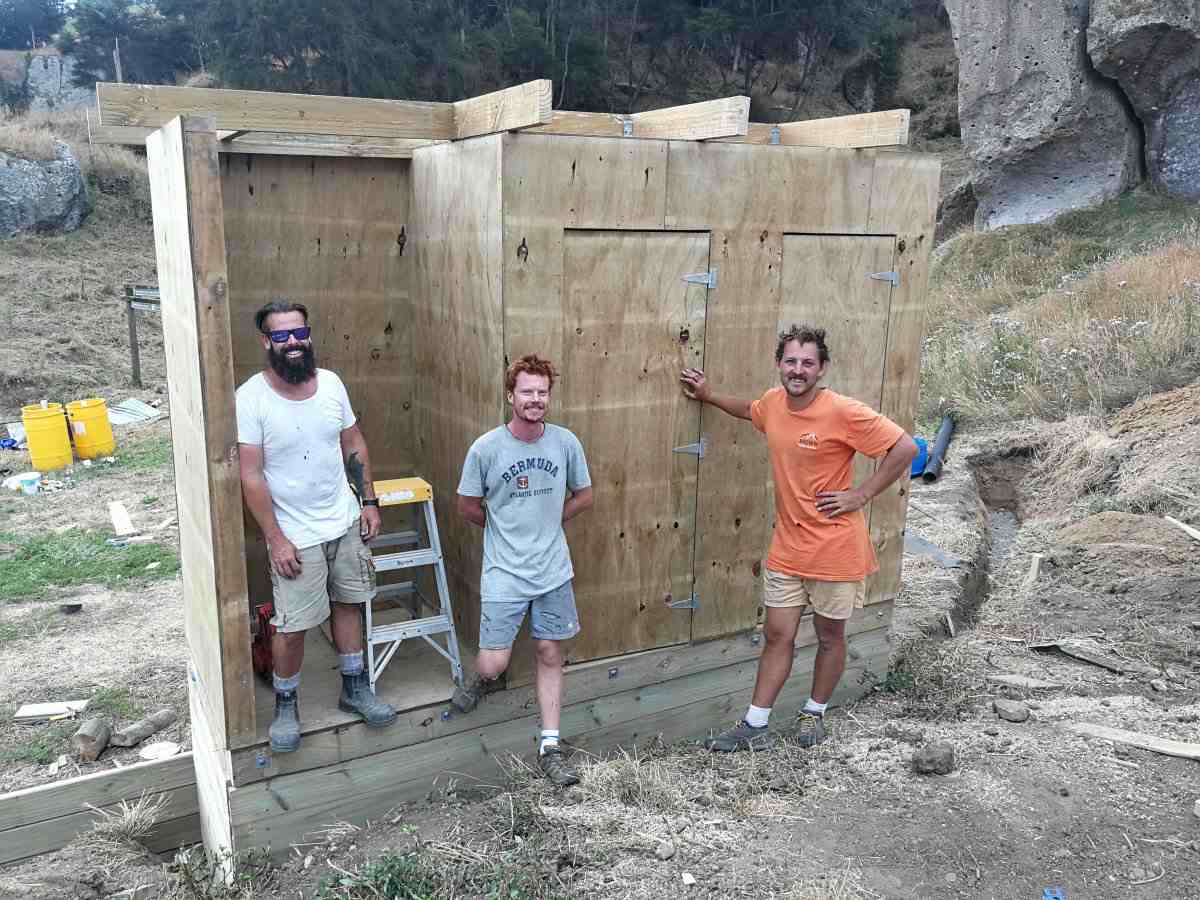Three builders in front of a plywood toilet block with cliffs in background.