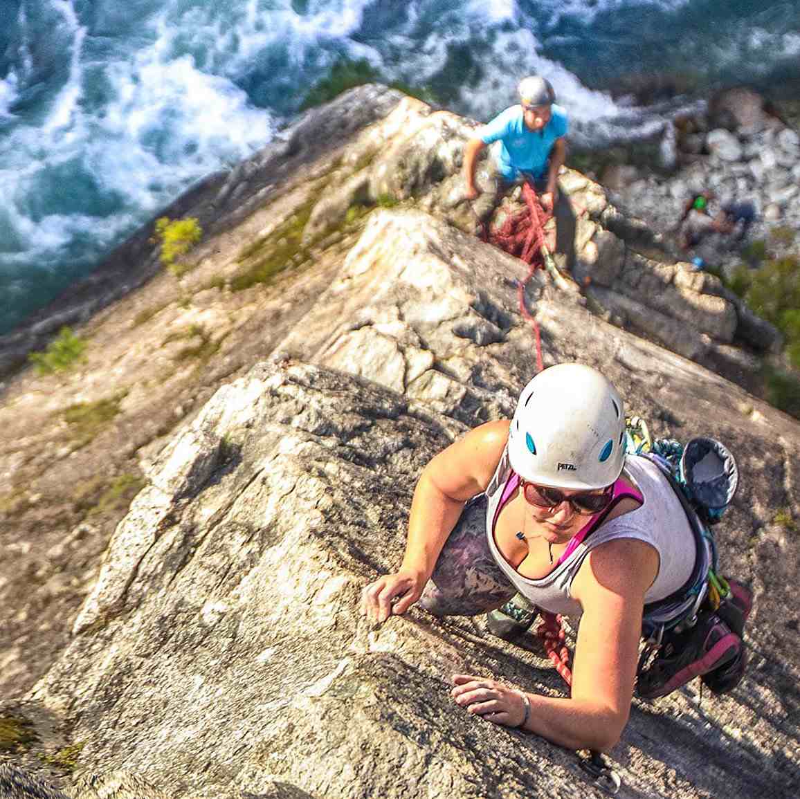 Looking down an arete at a female climber in white top and helmet, above blue river rapids,