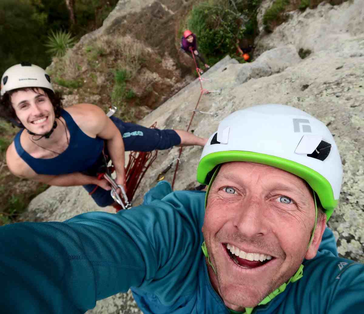 Close up selfie of smiling climber in white helmet, looking down at his climbing partners.