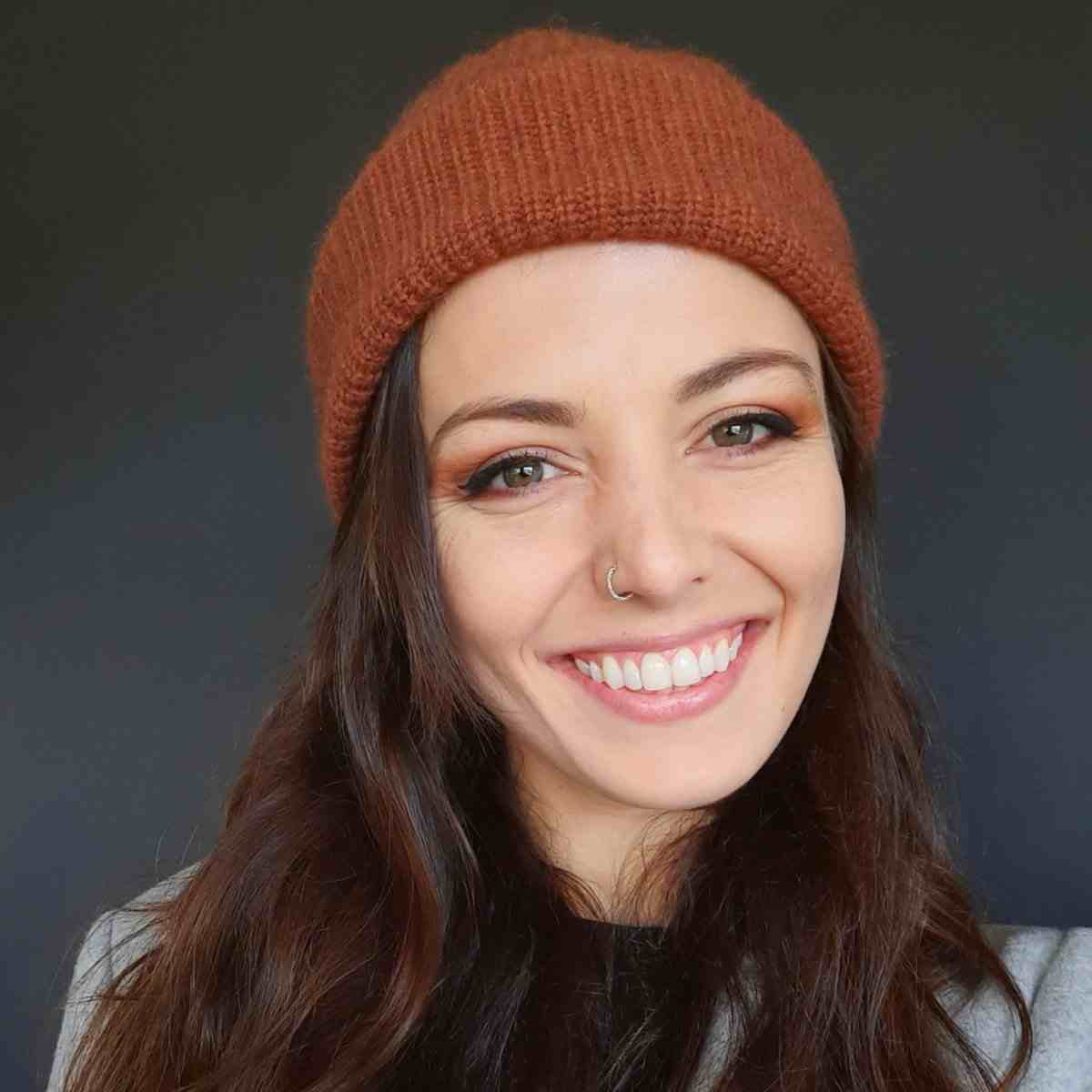 Smiling brunette woman with nose ring in read beanie hat.