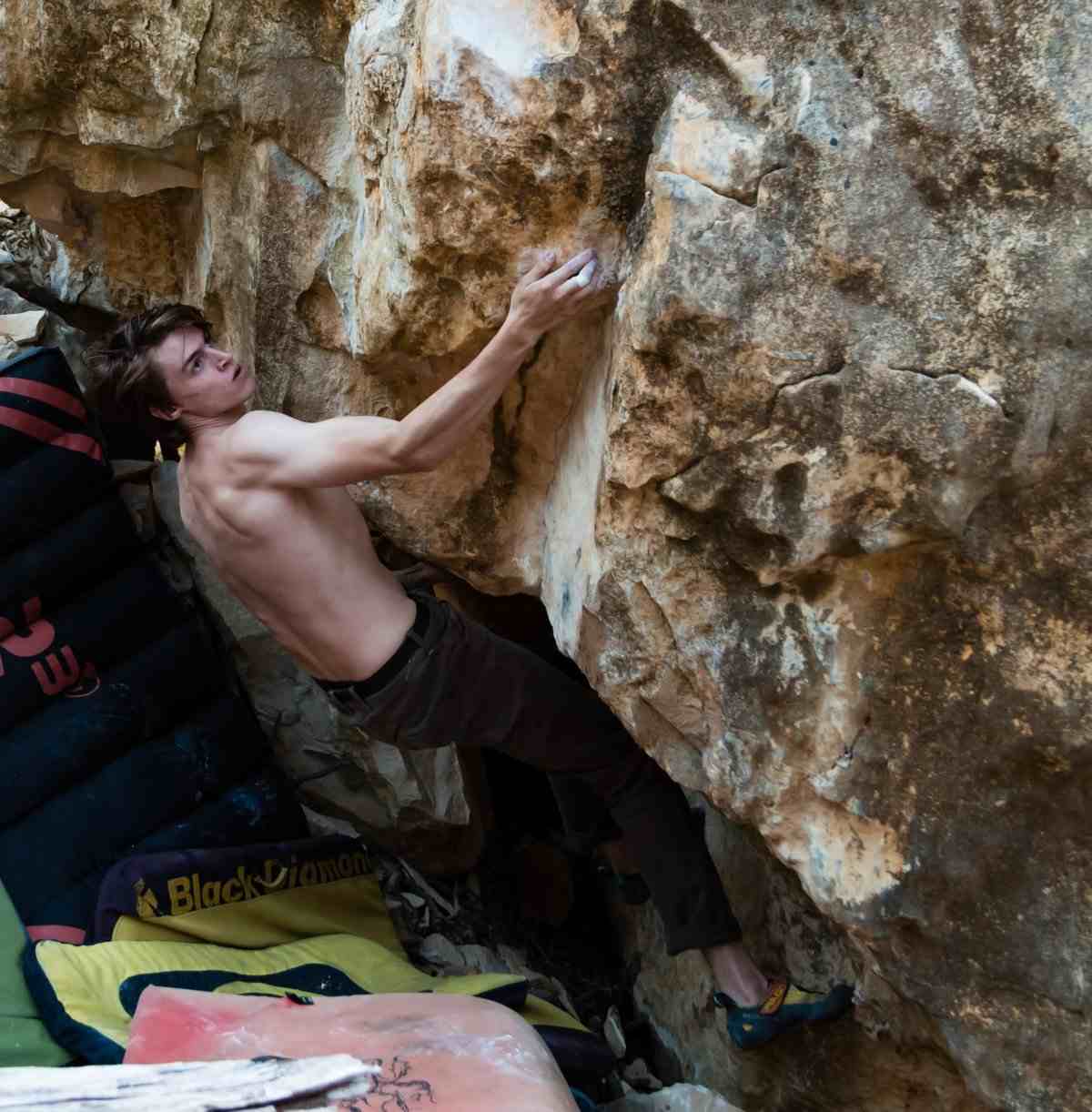 Shirtless climber on overhanging boulder with bouldering mats underneath