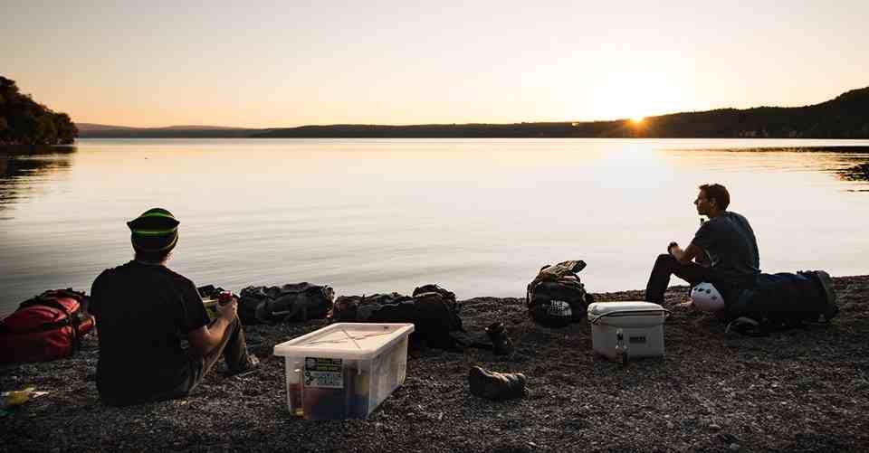 Two people sitting silhouetted on a gravel beach at sunset with camping gear.