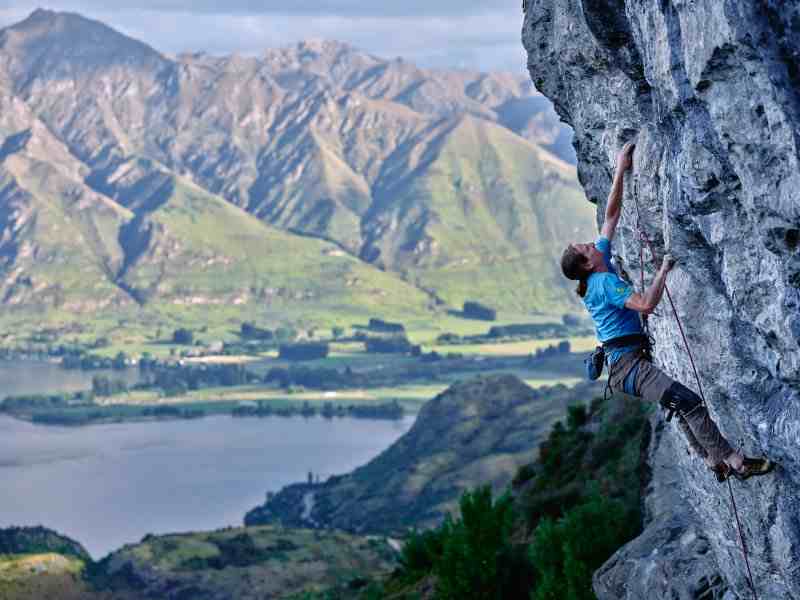 Climber in blue on steeply overhanging rock with mountains and lake in the background.